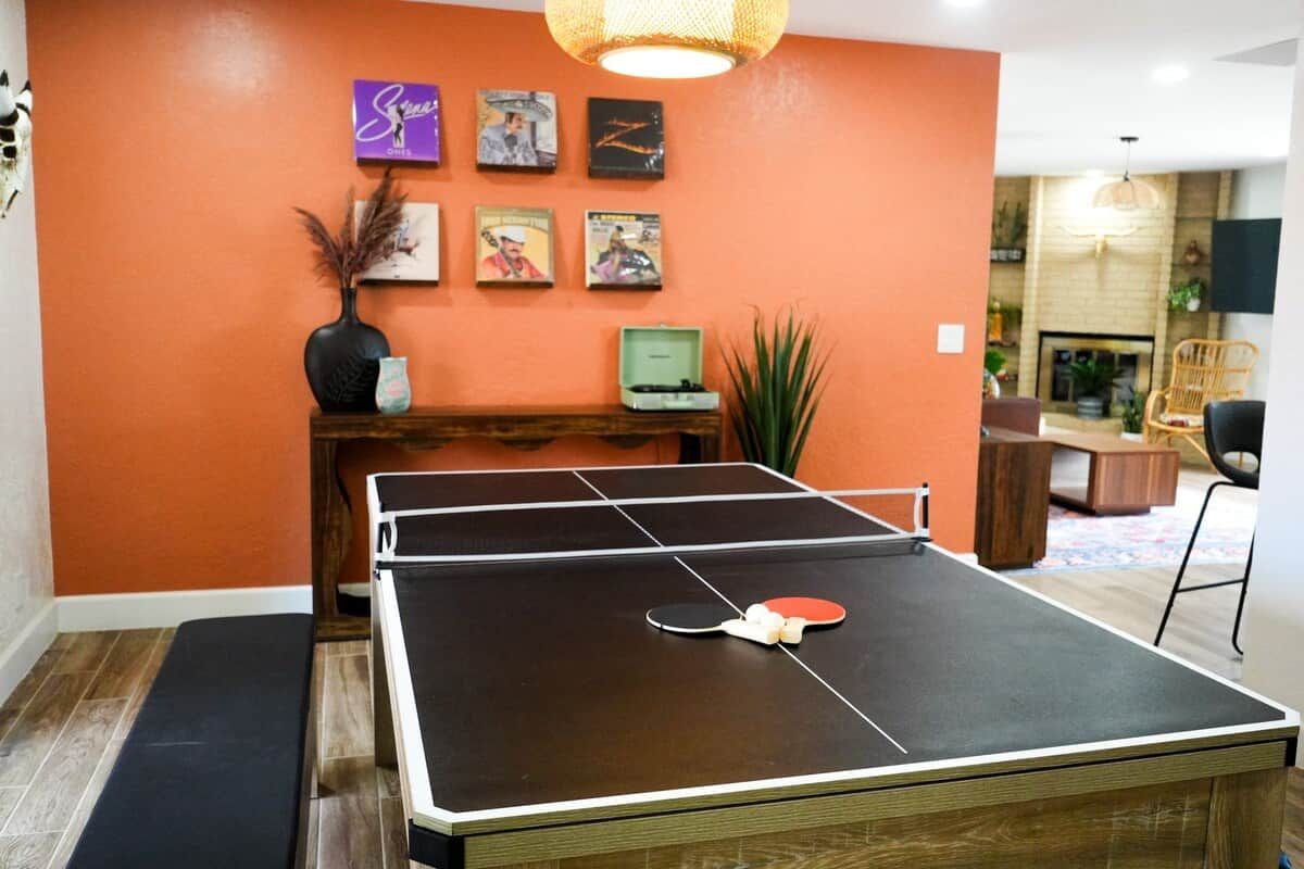Une table de ping-pong dans une pièce aux murs orange, avec un banc en bois et des tableaux encadrés accrochés au mur.