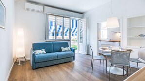 A modern living room with a blue sofa, a dining table with chairs, and a kitchen area visible through an open door. - Nice apartment for 4 guests with A/C, WIFI, TV and balcony (Cecina)