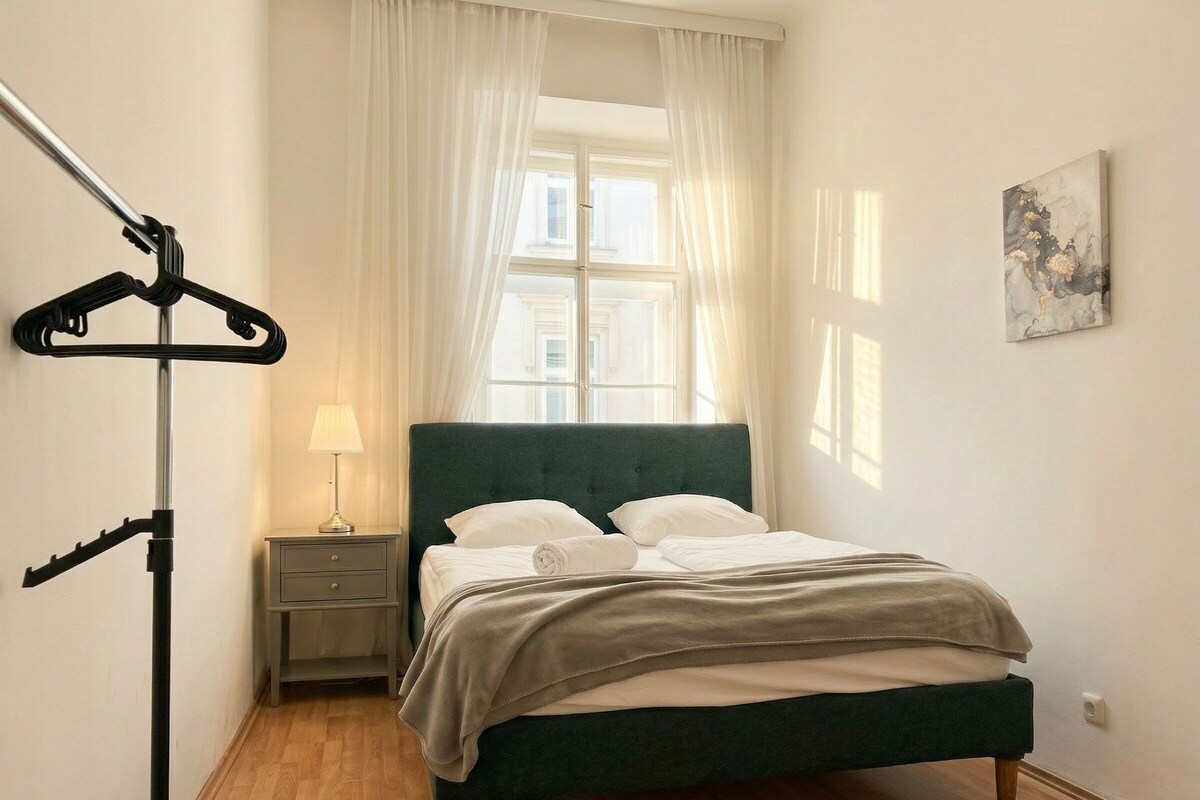 Ein ordentlich bezogenes Bett mit einem grünen Kopfteil, ein Nachttisch mit einer Lampe und ein Kleiderständer.