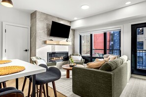 A modern living room with a fireplace, comfortable seating, a dining area, and large windows offering a view of the city. - Modern Rocky MTN Stay, Sleeps 8 with Pool +and  HotTub! (Canmore)