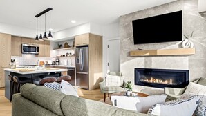 Uma sala de estar moderna com lareira, uma TV de tela plana acima dela e uma área de cozinha com armários de madeira e uma geladeira de aço inoxidável.