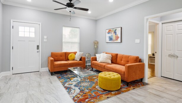 A living room with two orange sofas, a yellow ottoman, a glass coffee table, and a ceiling fan.