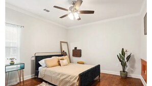 A bedroom with a bed, a ceiling fan, a bedside table, a chair, a window with blinds, a potted plant, and a wooden floor.