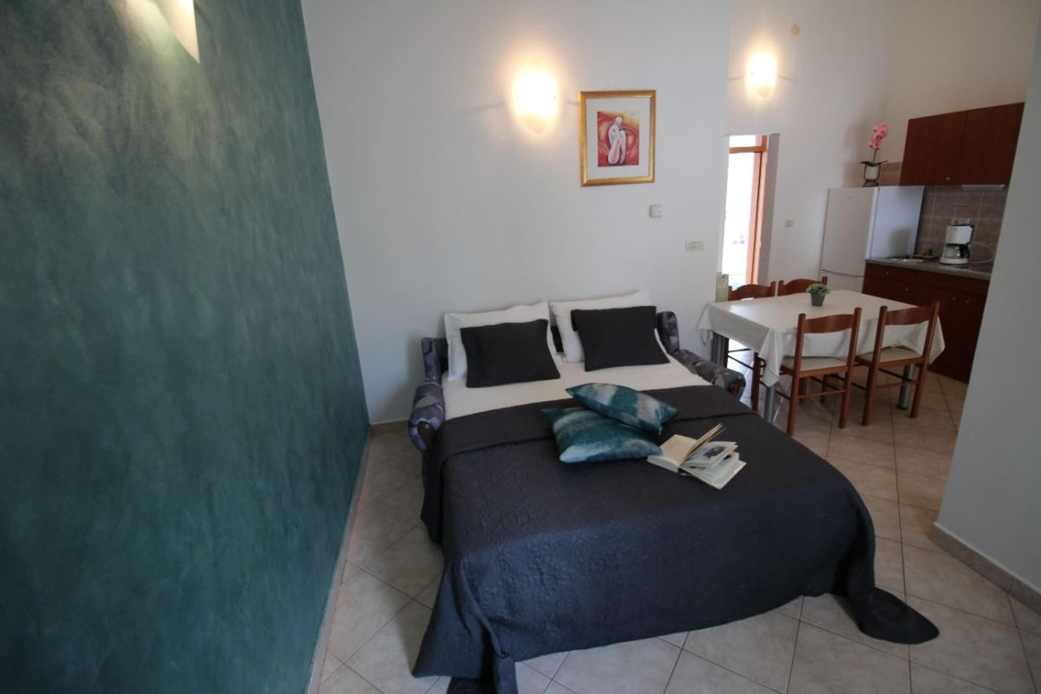 A bedroom with a bed, bedside tables, a small desk, and a chair. There is a kitchenette in the background.