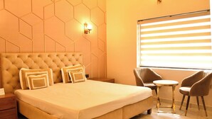 A hotel room with a large bed, a small round table, two chairs, a nightstand, and a window with blinds. - Shakti Resort (Behror)