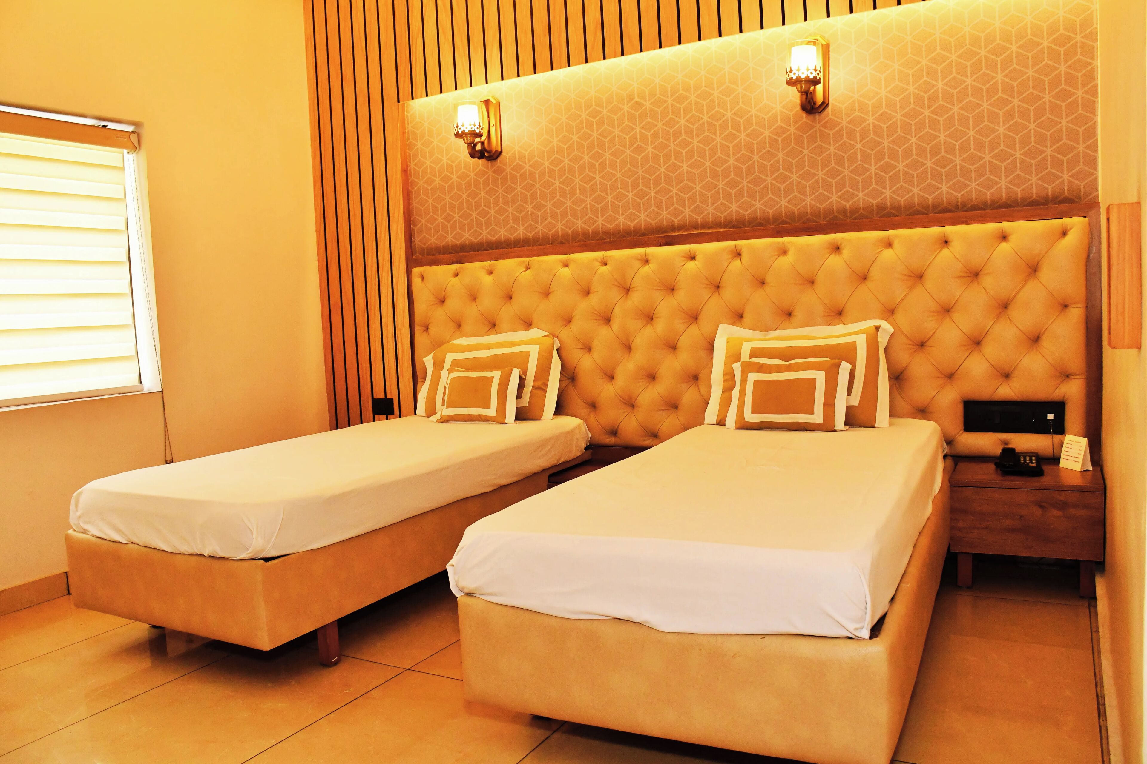 A hotel room with two single beds, a wooden headboard, a bedside table with a lamp, and a window with blinds.
