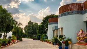 A paved pathway lined with potted plants leading to a building with a decorative fence and a statue. - Shakti Resort (Behror)