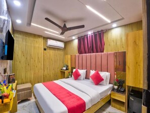 Ein Hotelzimmer mit einem Bett, einem Deckenventilator, einer Klimaanlage, einer Holzwand, einem kleinen Tisch mit einem Telefon und einem an der Wand montierten Fernseher.