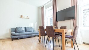 Een moderne woonkamer met een grijze bank, een houten eettafel met vier stoelen, een flatscreen televisie aan de muur en grote ramen met gordijnen.