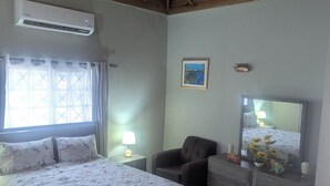 Ein Schlafzimmer mit Bett, Sessel, Kommode, Klimaanlage, einem Fenster mit Vorhängen und einem Bild an der Wand.