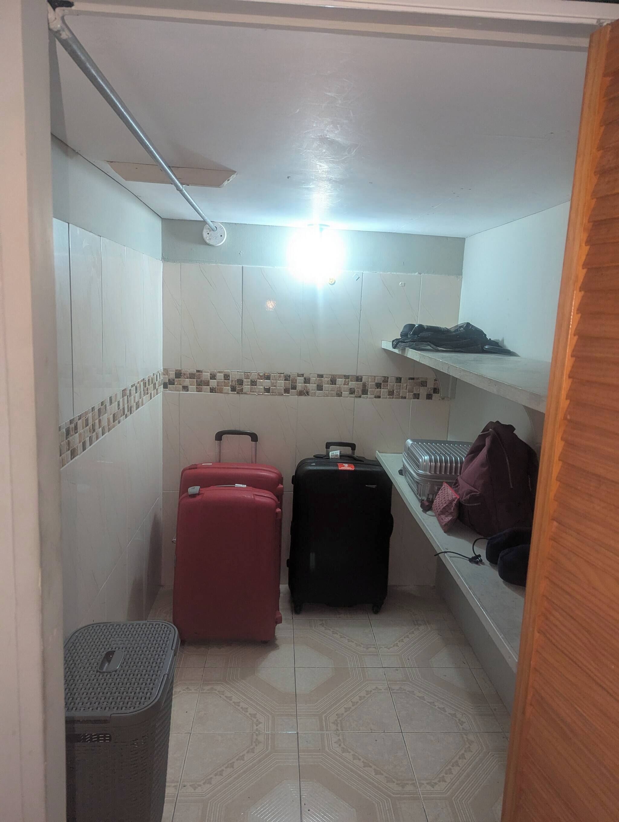 Une chambre d’hôtel de taille modeste, avec un sol carrelé, un ventilateur de plafond, une petite poubelle et deux valises.