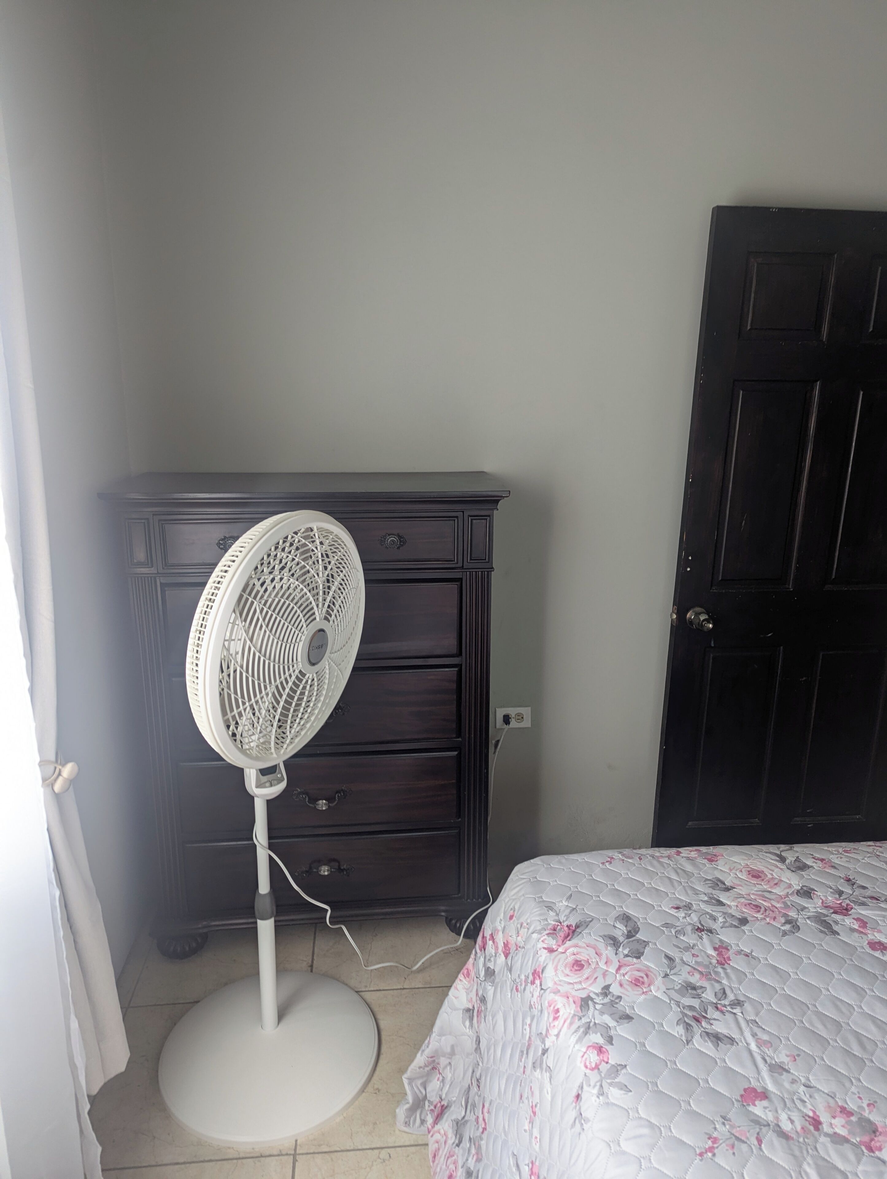 Une chambre avec une commode en bois foncé, un ventilateur blanc, un lit recouvert d’une courtepointe à motifs floraux et une porte en bois foncé fermée.