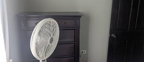 Une chambre avec une commode en bois foncé, un ventilateur blanc, un lit recouvert d’une courtepointe à motifs floraux et une porte en bois foncé fermée.