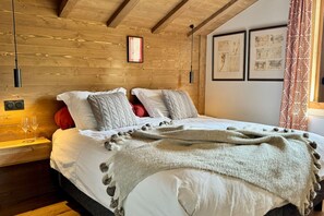 A bedroom with a wooden ceiling, a bed with white bedding and a grey blanket, two red pillows, and a bedside table with two glasses. - Megève 3BR 3BA · Spa, Pool, Fireplace (Megève)