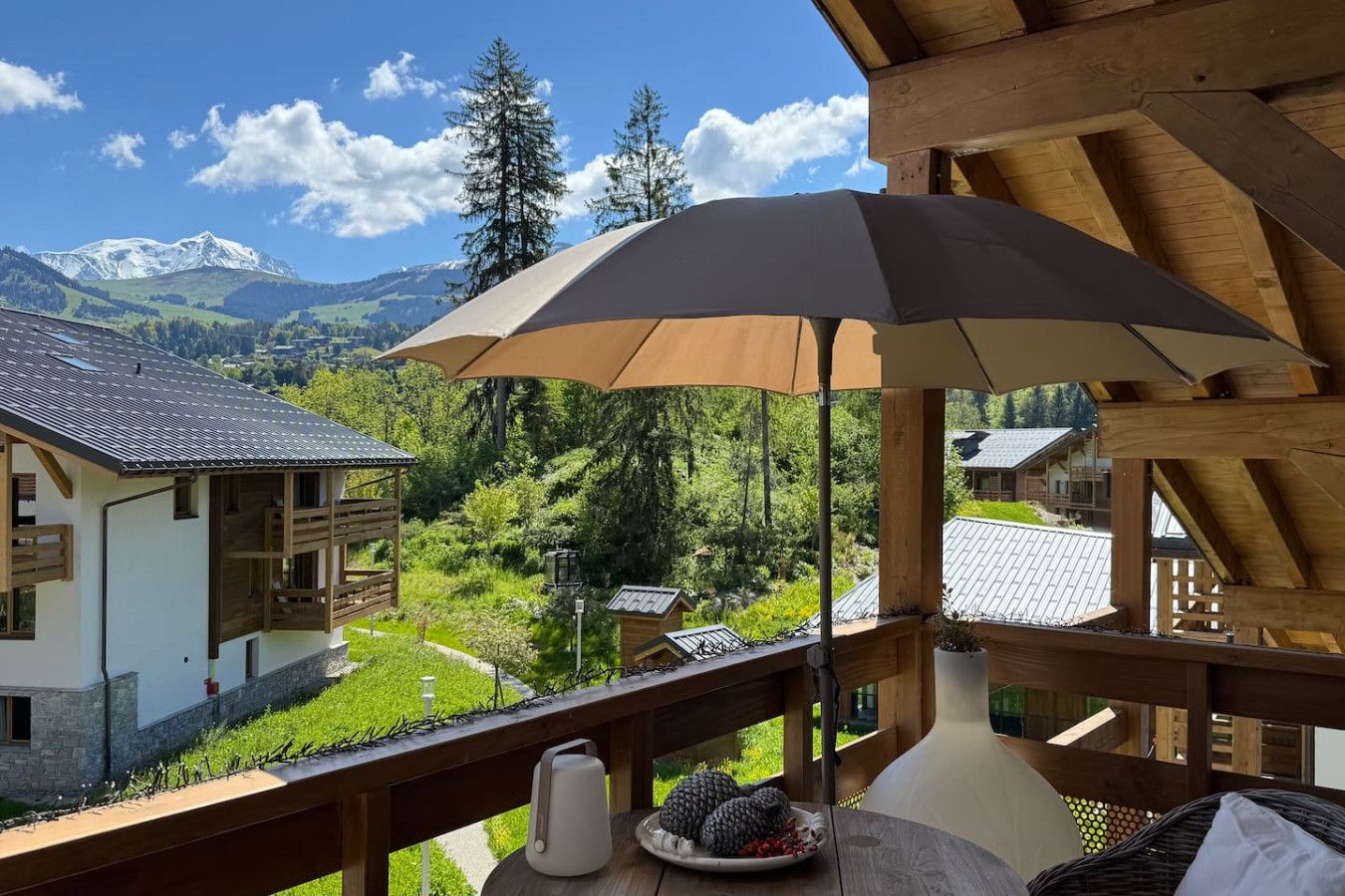 A wooden balcony with a view of a mountain landscape, a table with a plate of berries, and a large umbrella.