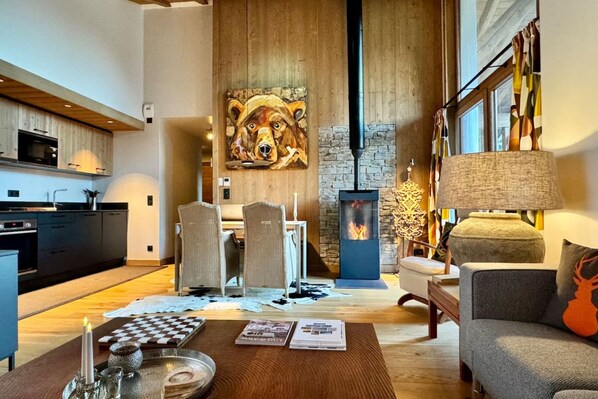 A modern living room with a fireplace, a dining area with chairs, a sofa, and a coffee table with books and a candle. - Megève 3BR 3BA · Spa, Pool, Fireplace (Megève)