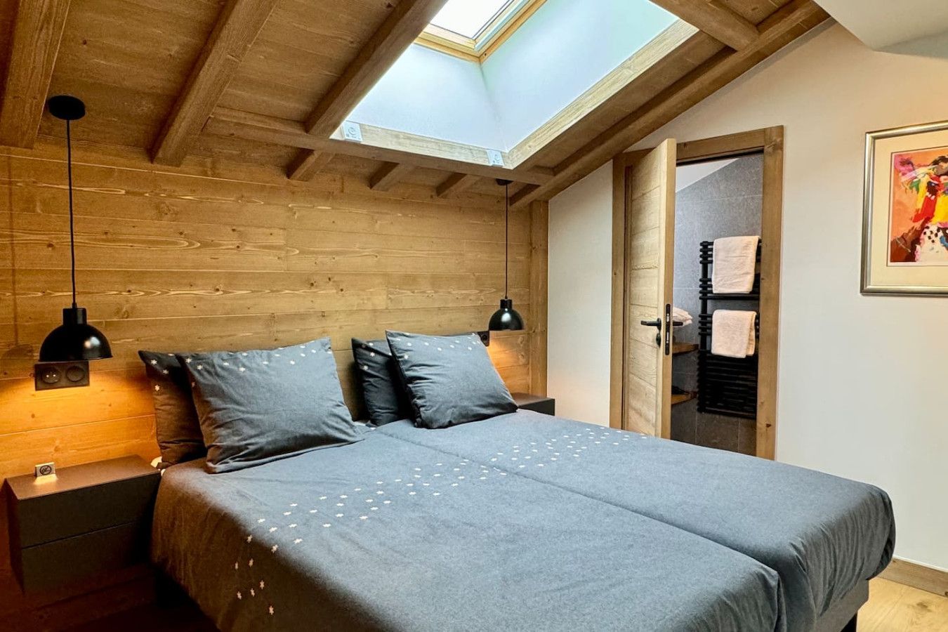 A bedroom with a skylight, wooden ceiling beams, a bed with grey bedding, two bedside lamps, and a door leading to a bathroom with towels.