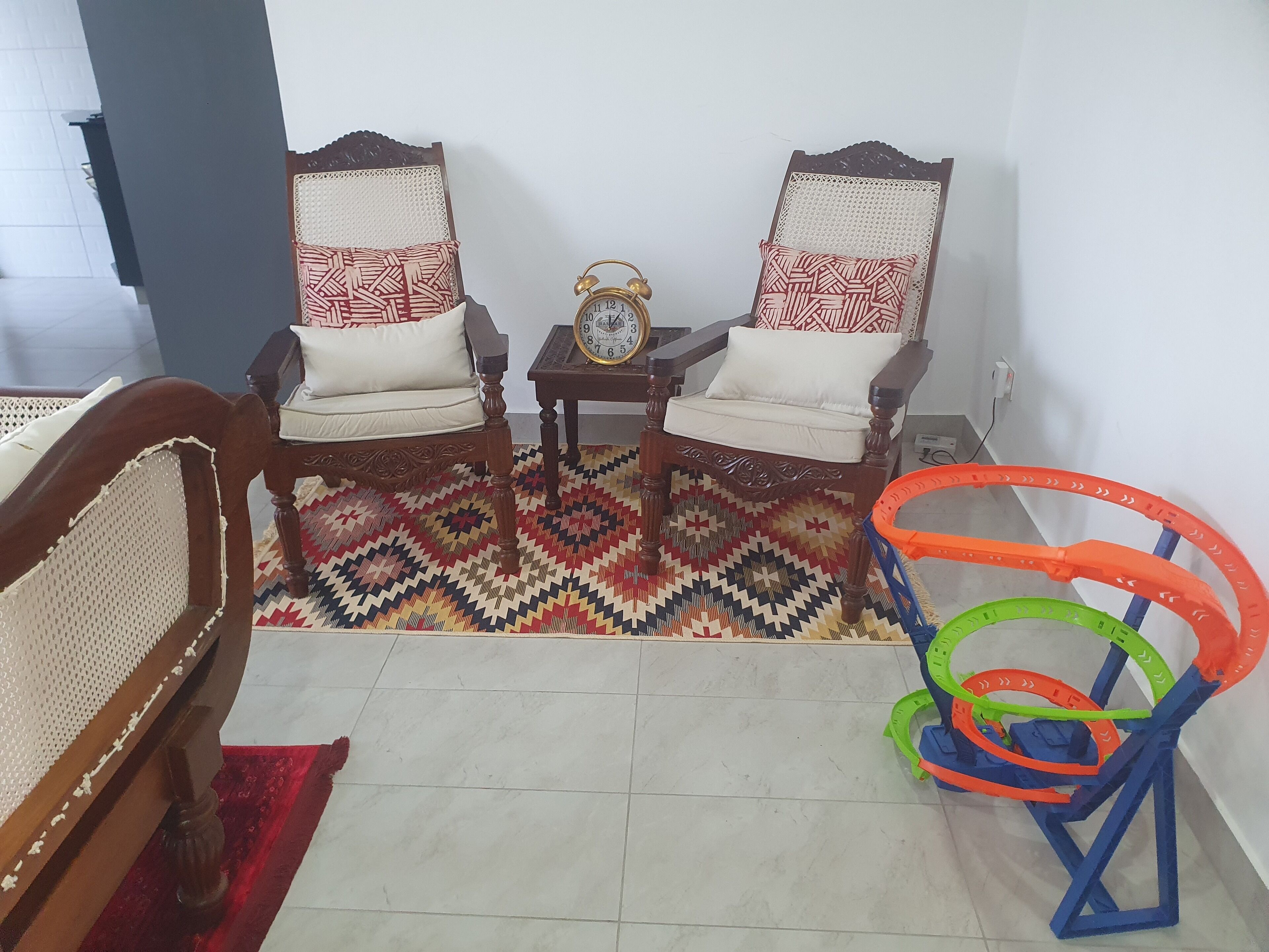 A living room with two wooden chairs with white cushions, a small table with a clock, and a colorful woven rug.