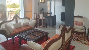 A living room with a sofa, a coffee table, and a patterned rug. There is a kitchen area with a counter and a hanging light fixture. - Luxury Mombasa Apartment with Ocean and City Views (Mombasa)