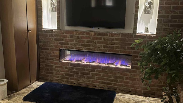 A living room with a TV mounted on a brick wall, a fireplace below, and a sofa with a remote control on it. - Wonky 2– A Cosy Hideaway in the Heart of Cheshire (Macclesfield)
