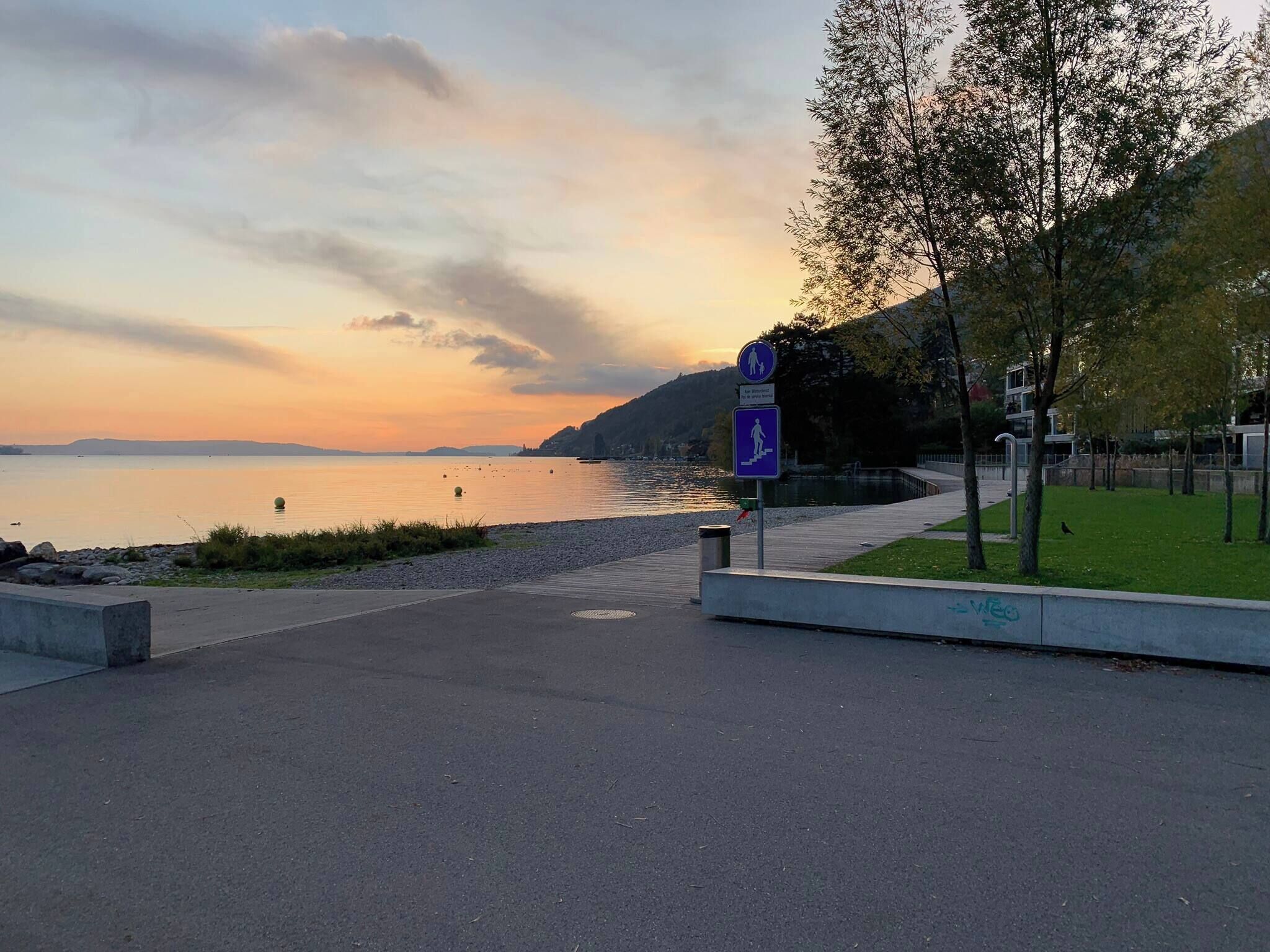 Eine Uferpromenade mit Fußweg, einem Schild, das einen Wanderweg weist, und ruhigem Wasser bei Sonnenuntergang.