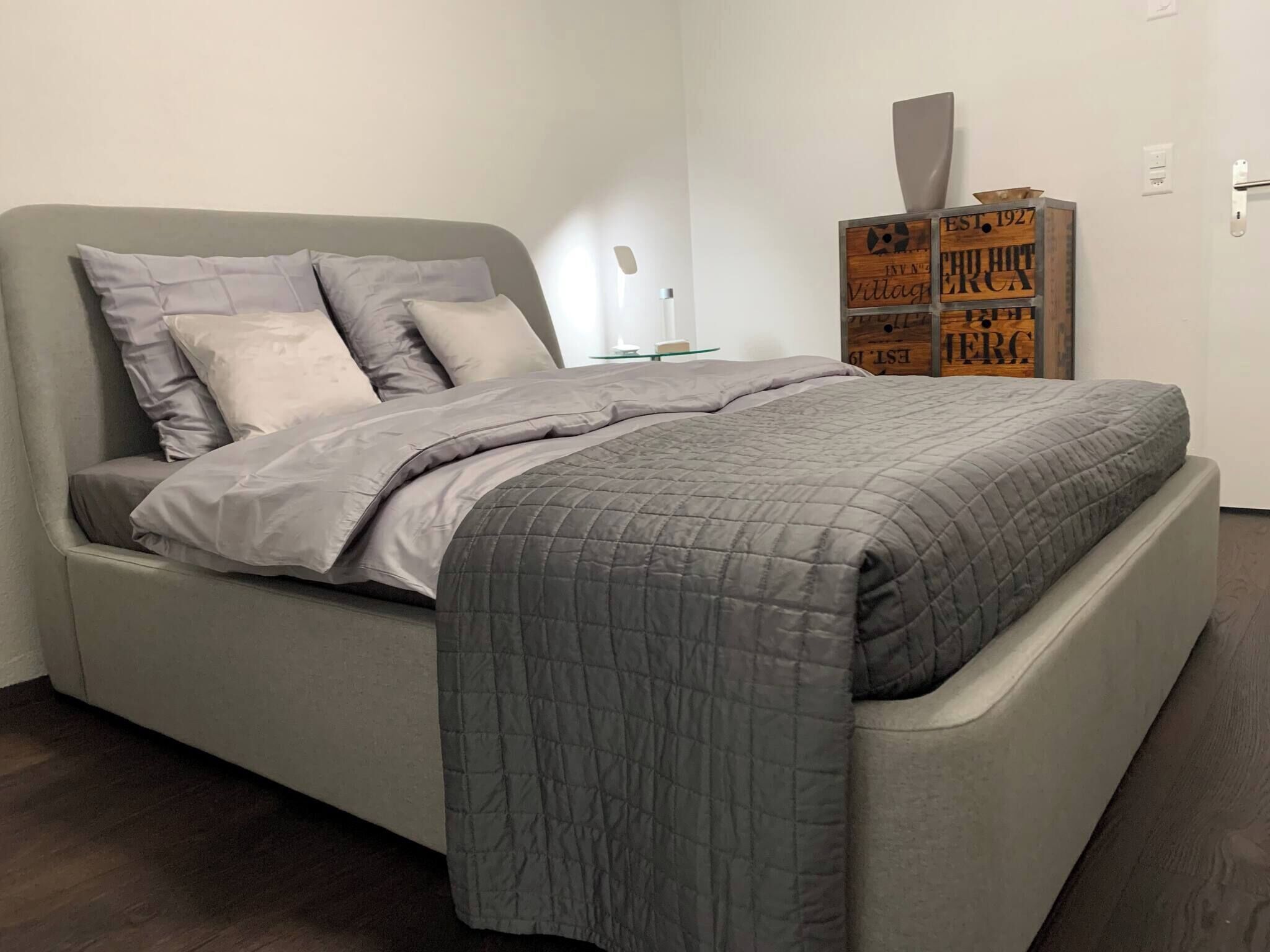 Une chambre moderne avec un lit gris, une couverture matelassée grise et une table de chevet avec une lampe. Des caisses en bois sont empilées contre le mur.