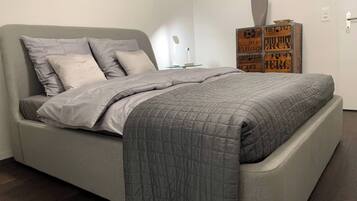 Une chambre moderne avec un lit gris, une couverture matelassée grise et une table de chevet avec une lampe. Des caisses en bois sont empilées contre le mur.