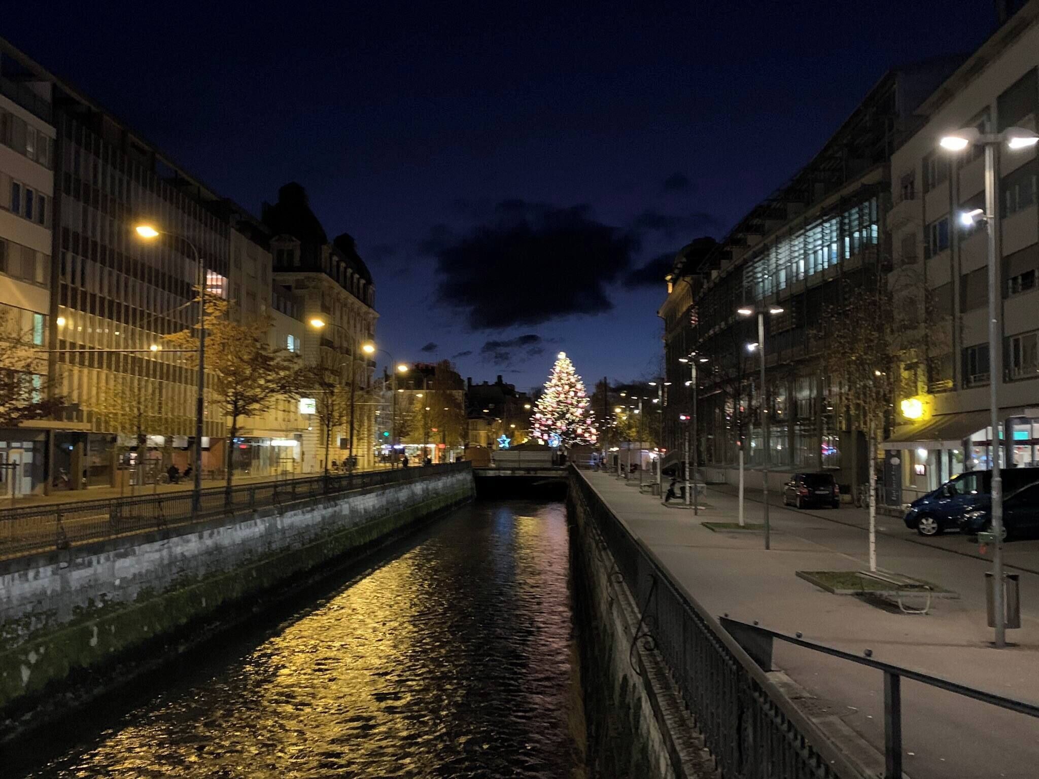 Un paisaje urbano nocturno con un árbol de Navidad iluminado a lo lejos, un canal y edificios modernos.