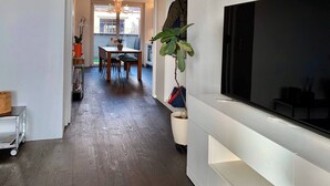 Una sala de estar moderna con televisor de pantalla plana, piso de madera, un área de comedor con mesa y sillas, y una planta en maceta.