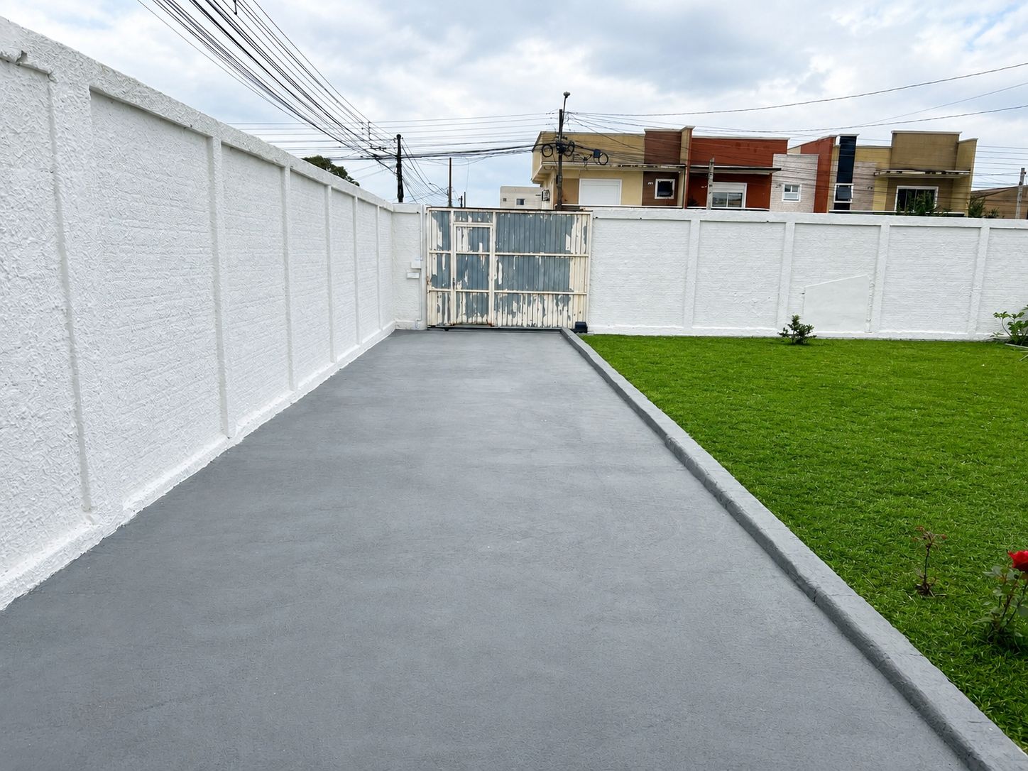 A concrete pathway leading to a gated entrance, surrounded by a white wall and a grassy area.