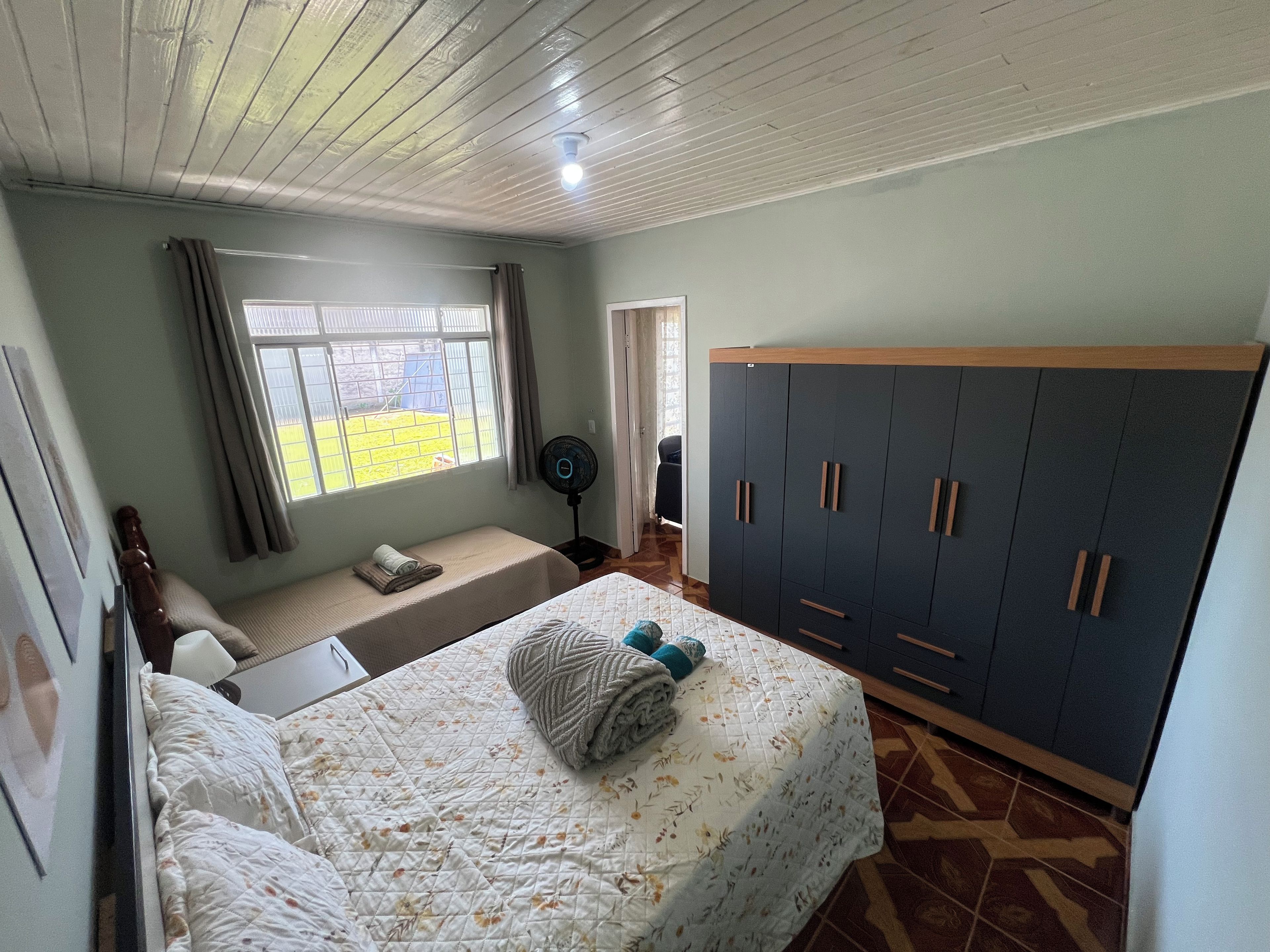A bedroom with a bed, a wardrobe, a window with curtains, and a fan.