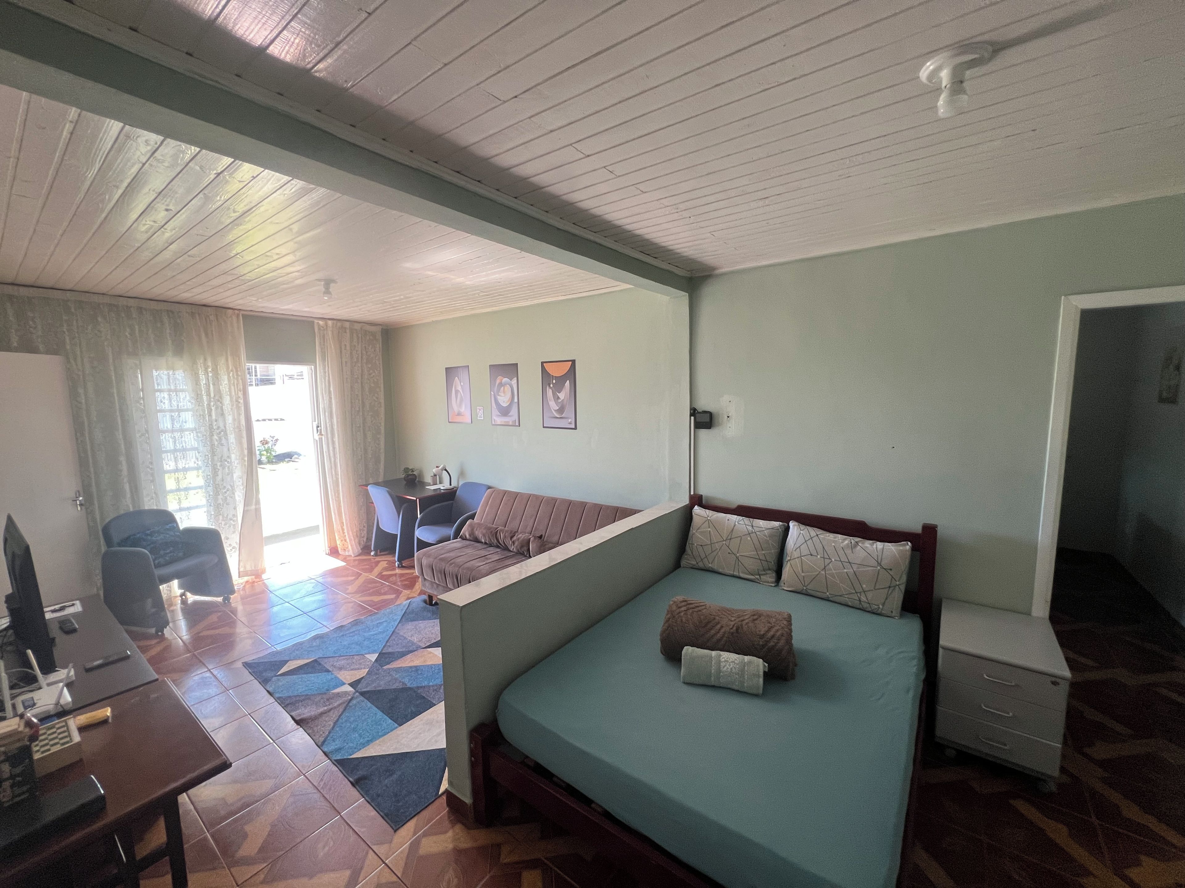 A room with a bed, a desk, a chair, and a small table. There is a window with curtains, a wall with framed pictures, and a ceiling fan.
