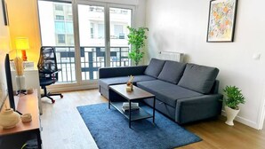 A modern living room with a grey sofa, a coffee table, and a blue rug. There is a large window with a view of buildings outside. - L'Ensoleillé Levallois – Apartment near metro (Levallois-Perret)