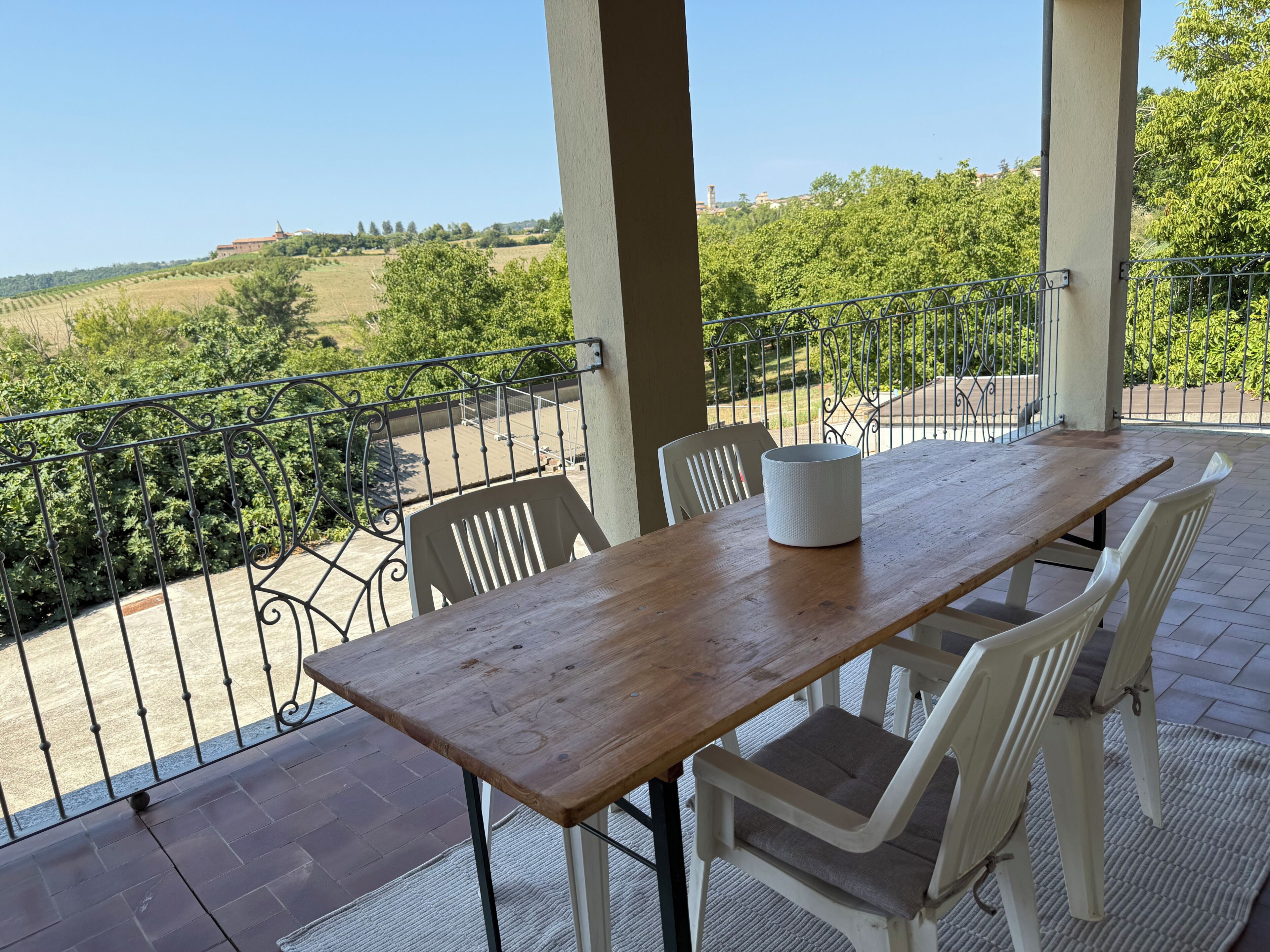 A wooden table with chairs on a balcony with a metal railing, overlooking a rural landscape.