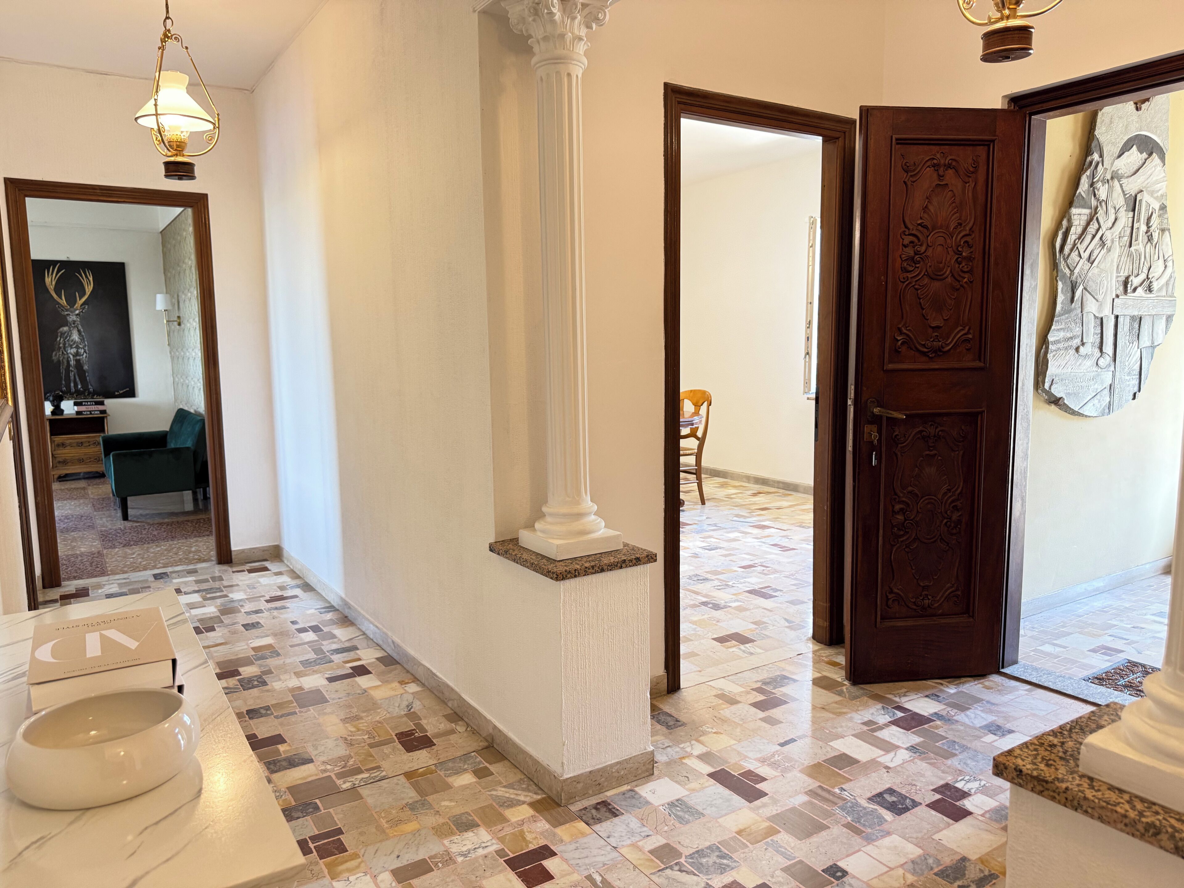 Un pasillo con piso de baldosas, un pilar blanco, una puerta de madera con tallas intrincadas y una obra de arte enmarcada en la pared.