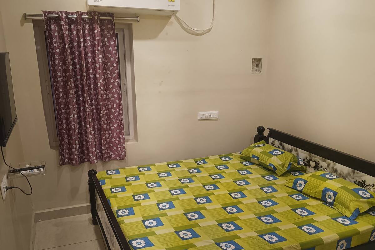 Um quarto com cama, ventilador de teto, ar condicionado, janela com cortinas e televisão fixada na parede.