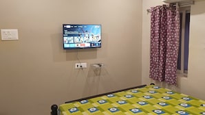 A bedroom with a bed, a ceiling fan, a TV mounted on the wall, a window with curtains, and a power outlet. - Hotel Homestay Bhimavaram (Bhimavaram)