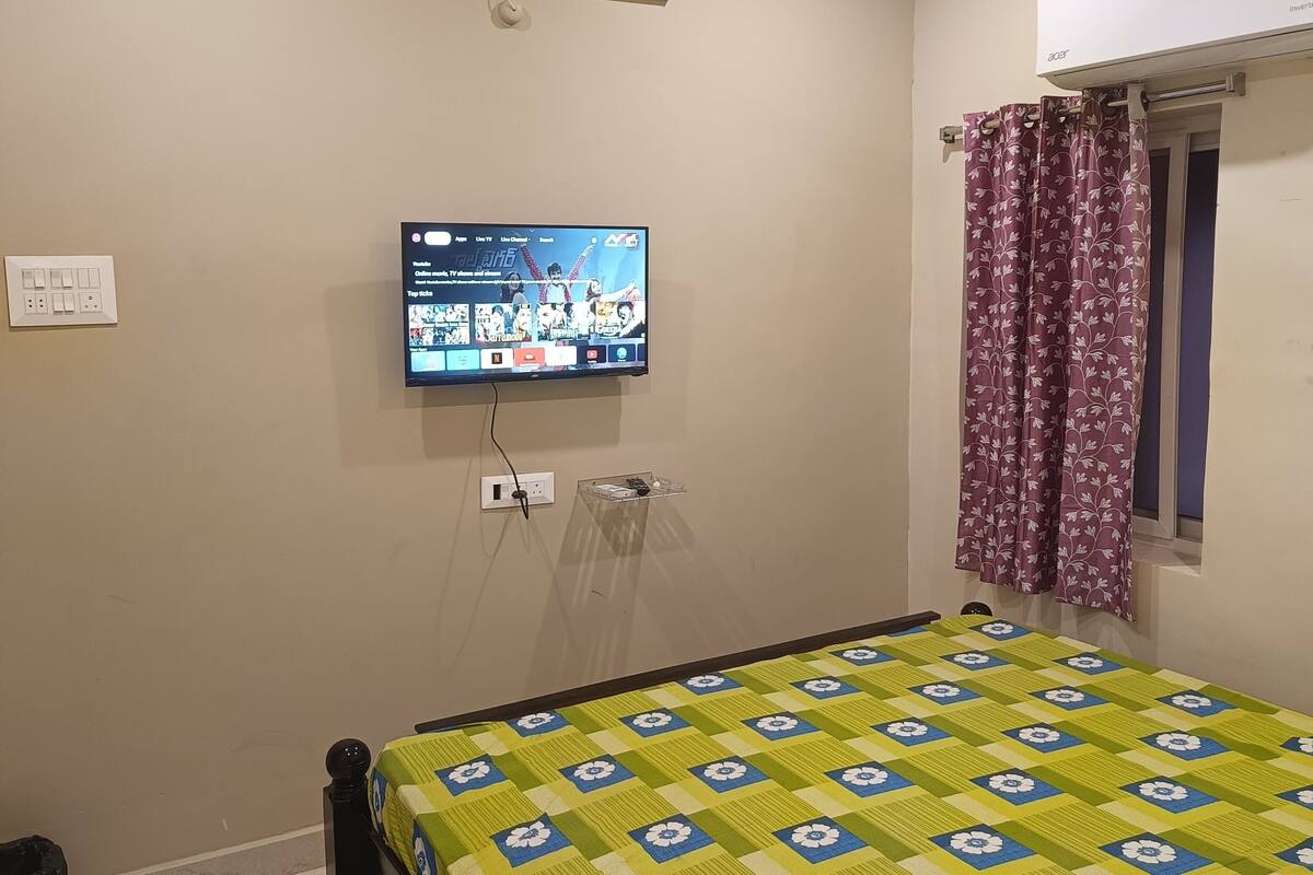 Quarto com cama, ventilador de teto, TV fixada na parede, janela com cortinas e tomada.