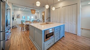 A kitchen with a large island, stainless steel appliances, and a dining area with wooden chairs. - Steps to Beach • Pool + Fire Pit + Golf Cart Boardwalk Access | Southern Cross (Port Aransas)
