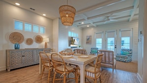 A dining area with a white table, wicker chairs, and a hanging basket light fixture. - Steps to Beach • Pool + Fire Pit + Golf Cart Boardwalk Access | Southern Cross (Port Aransas)
