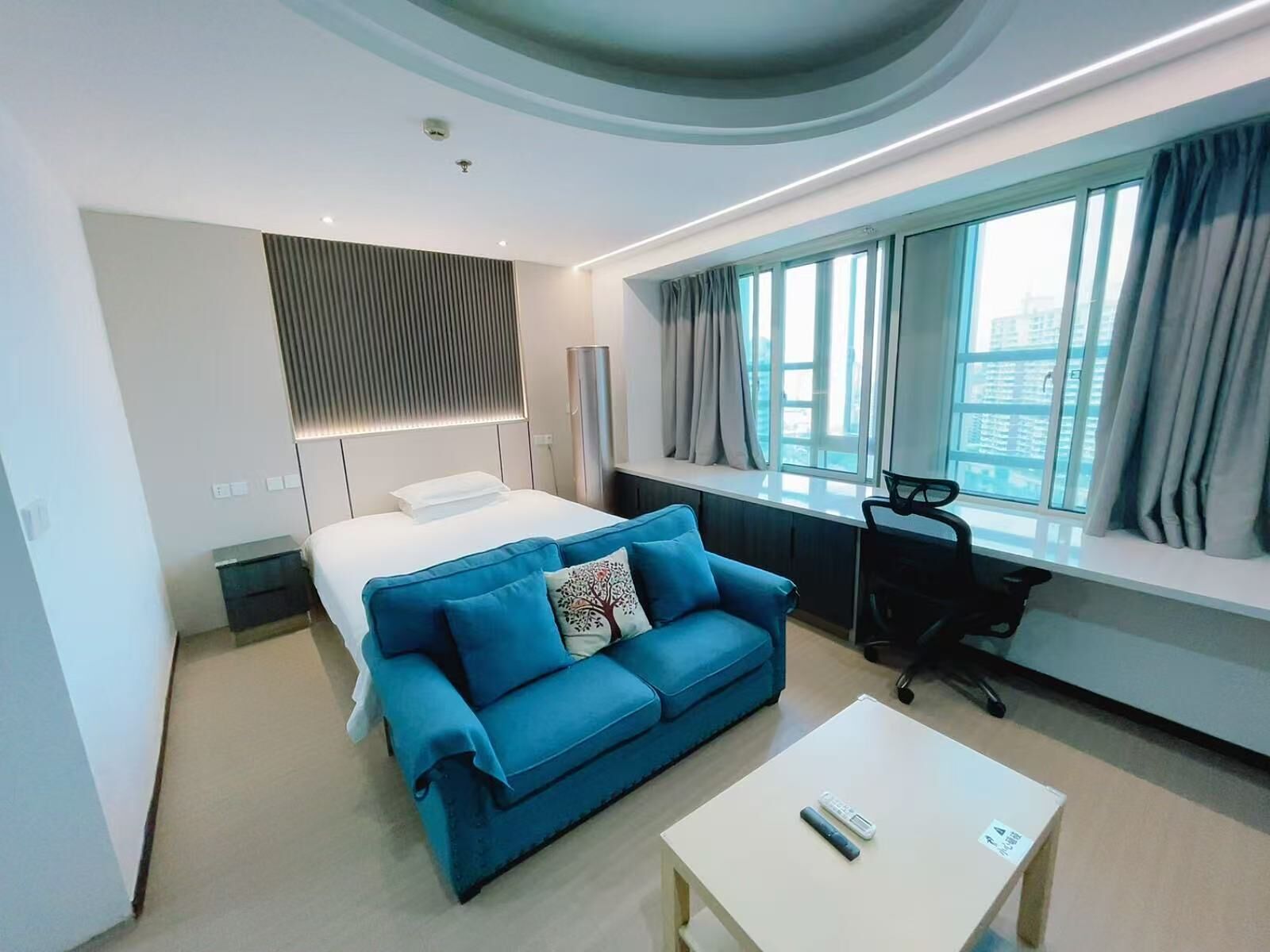 A modern hotel room with a bed, a blue sofa, a desk, and a large window with curtains.