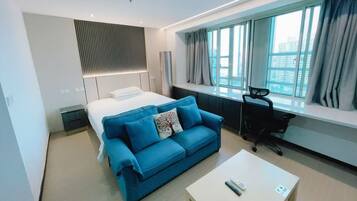 A modern hotel room with a bed, a blue sofa, a desk, and a large window with curtains.