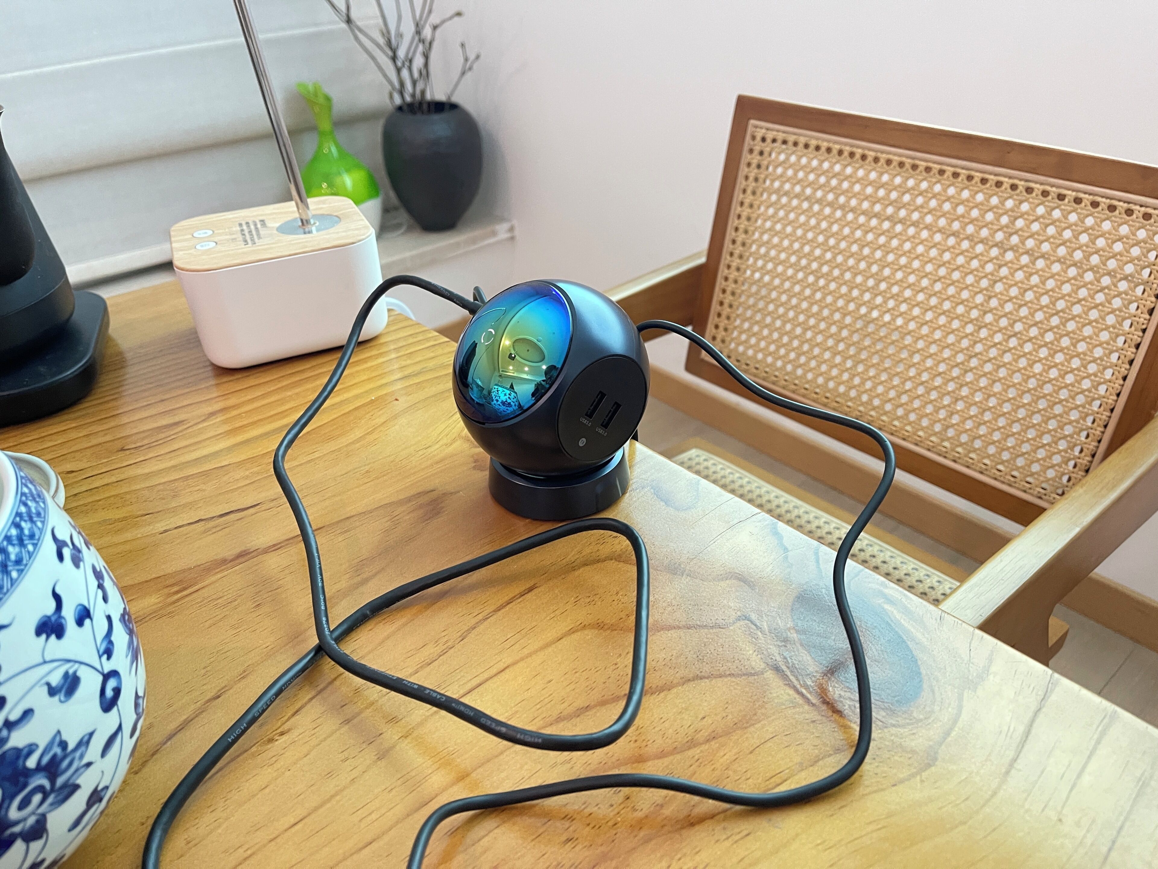 A round, black speaker with a visible logo on its front, connected by a cable to a wooden table.