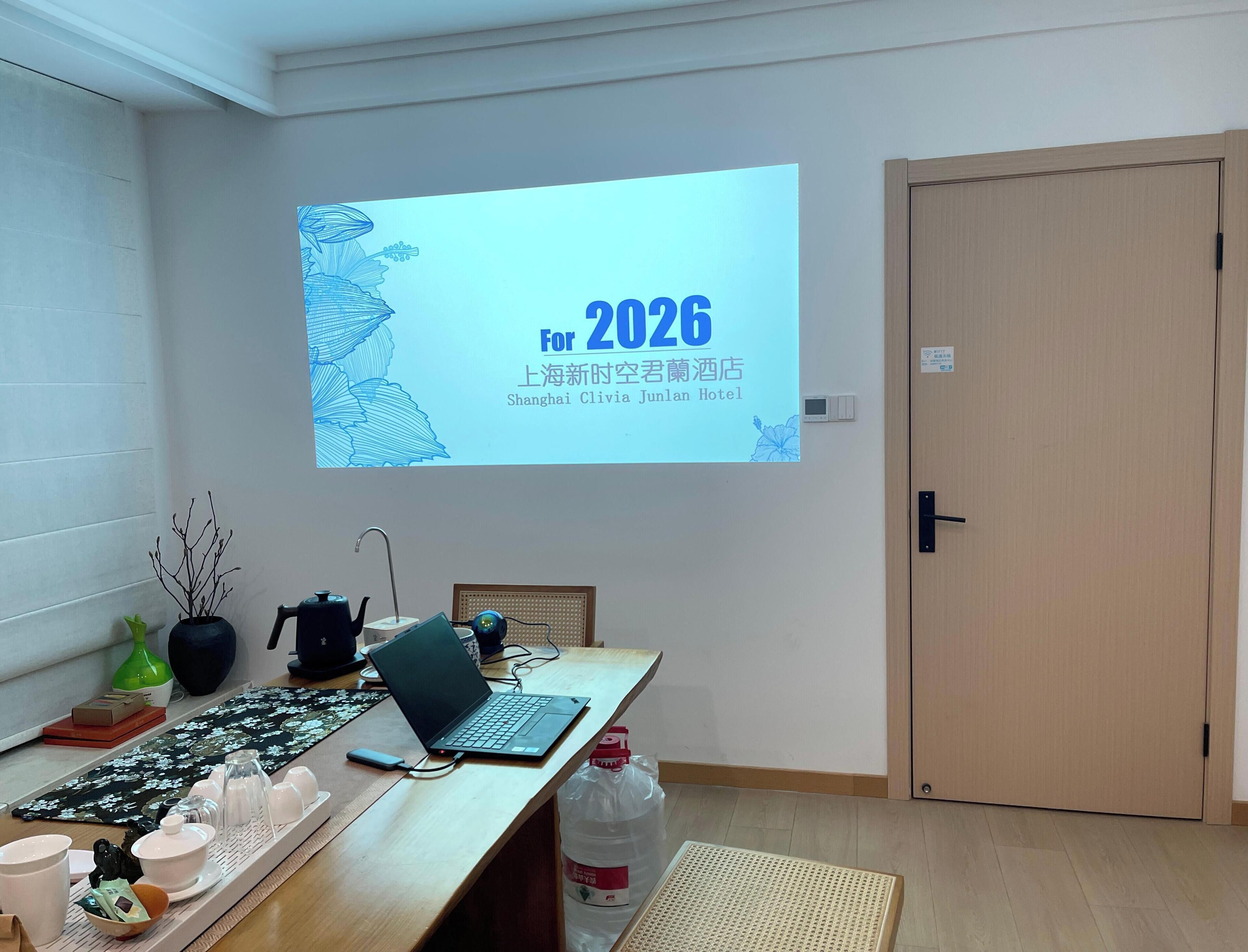 Une chambre d’hôtel équipée d’un écran de projection affichant les mots « 2026 » et « Shanghai Clivia Junlan Hotel ». On y trouve un bureau en bois avec un ordinateur portable, un plateau contenant du matériel à thé et une bouteille d’eau.