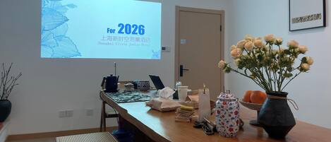 A hotel room with a wooden desk, a projector screen displaying a message for 2026, a vase with flowers, and a framed picture on the wall.