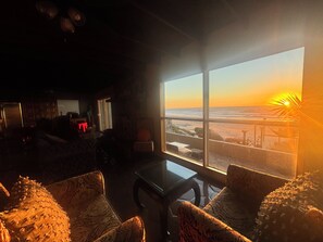 Ein Zimmer mit großem Fenster und Blick auf einen Strand bei Sonnenuntergang, mit einer Couch und einem Tisch.