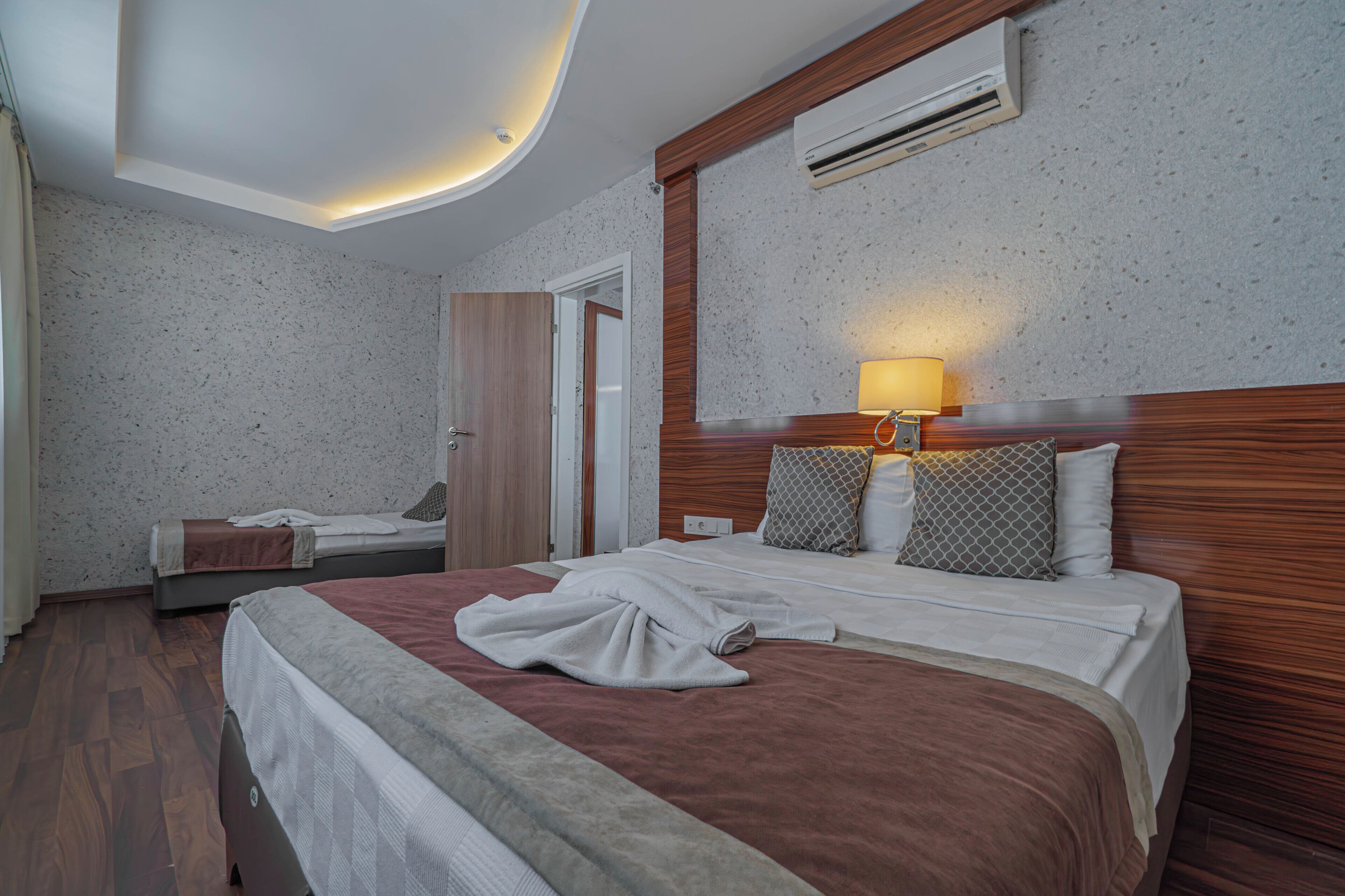 A hotel room with a large bed, a smaller bed, a wooden headboard, a wall-mounted air conditioner, and a ceiling light.