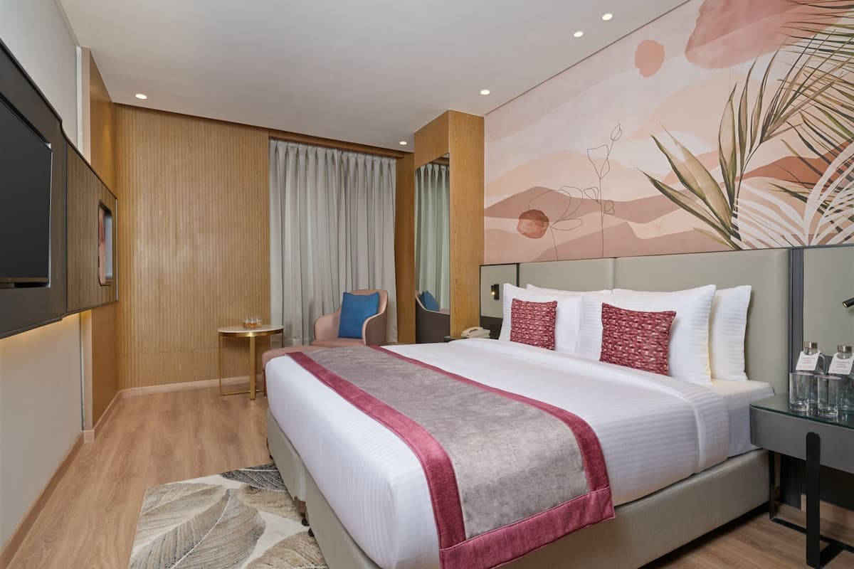 A hotel room with a large bed, a desk, a chair, a small table, and a wall with a plant and abstract design.