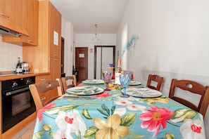 Un coin repas avec une table dressée pour quatre personnes, des chaises en bois, un espace cuisine équipé d’une cuisinière et de placards, et une porte avec une pancarte.