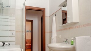 Uma casa de banho com uma pia branca, um espelho, chuveiro e sanita.